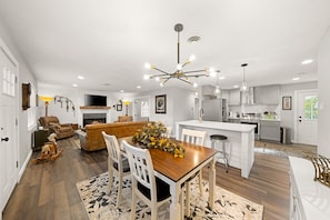 A spacious living area with a wooden dining table, white chairs, a sofa, and a fireplace.