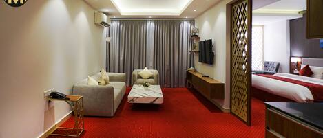 Habitación de hotel con sofá, sillón, una pequeña mesa con un florero, cama, televisor y un separador decorativo.