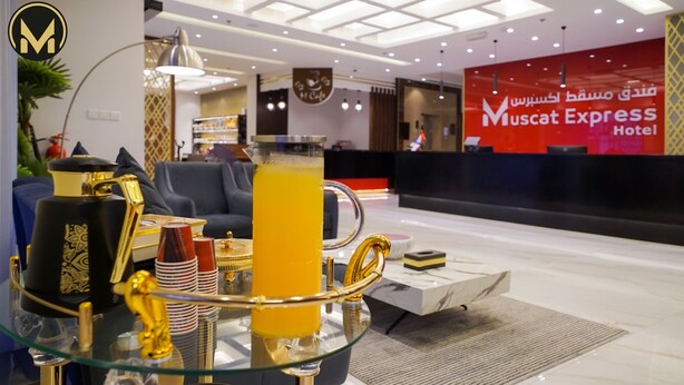 A reception area with a glass table displaying a golden saxophone, a red and white striped cup, and a glass of orange juice. There is a red sign with Arabic text and a logo in the background. - Muscat Express Hotel (Muscat)