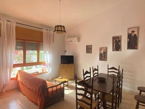 A cozy living room with a sofa, dining table, and chairs. There is a TV on a stand and a window with curtains.