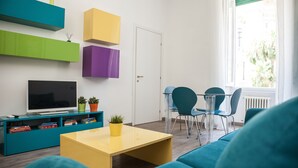 Un salon moderne avec des canapés de couleur turquoise, une table basse jaune et un téléviseur à écran plat posé sur un meuble turquoise. On y trouve également des meubles de rangement colorés fixés au mur, ainsi qu’un coin repas avec une table et des chaises.