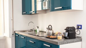 Une cuisine moderne avec des armoires de couleur turquoise, un évier en acier inoxydable, une bouilloire, une machine à café et un verre de jus d’orange posé sur le comptoir.