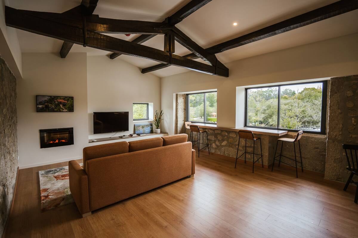 A spacious living area with a wooden floor, a brown sofa, a flat-screen TV, a fireplace, and large windows offering a view of trees.
