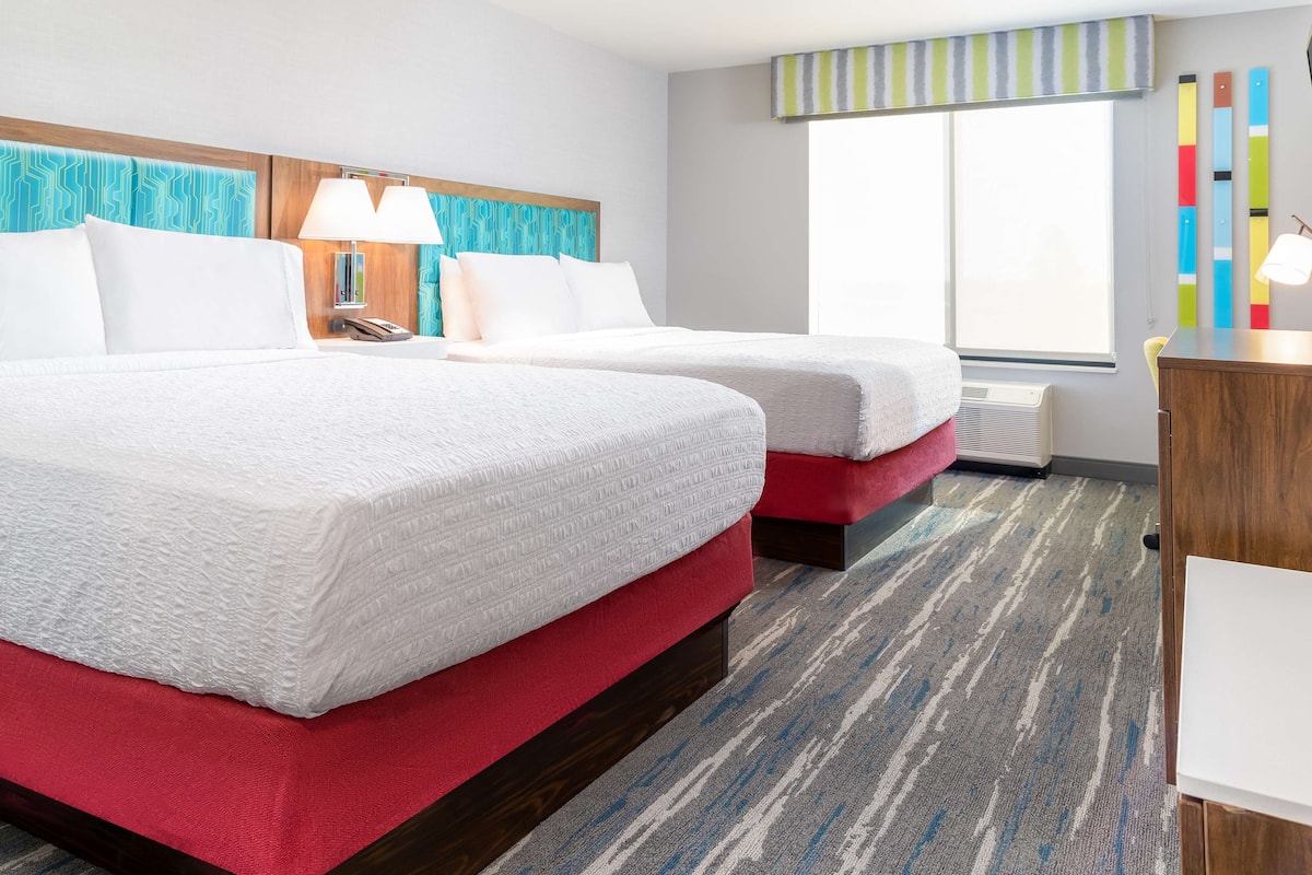 A hotel room with two beds, a wooden headboard, a striped window valance, a large window, and a carpeted floor.