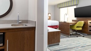 A hotel room with a wooden desk, a modern chair, a bed with a red blanket, a large mirror, and a flat-screen TV.