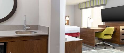 A hotel room with a wooden desk, a modern chair, a bed with a red blanket, a large mirror, and a flat-screen TV.