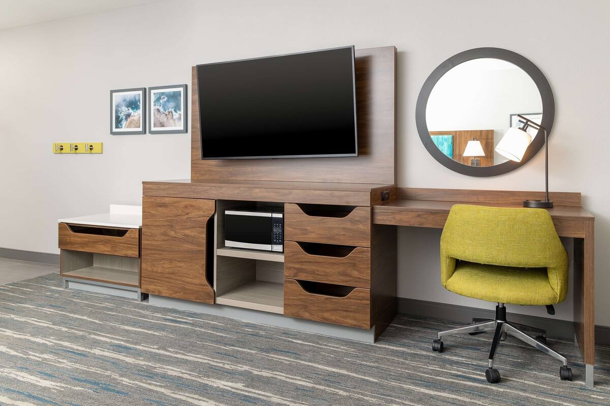 A modern hotel room with a wooden desk, a flat-screen TV, a round mirror, and a yellow office chair.