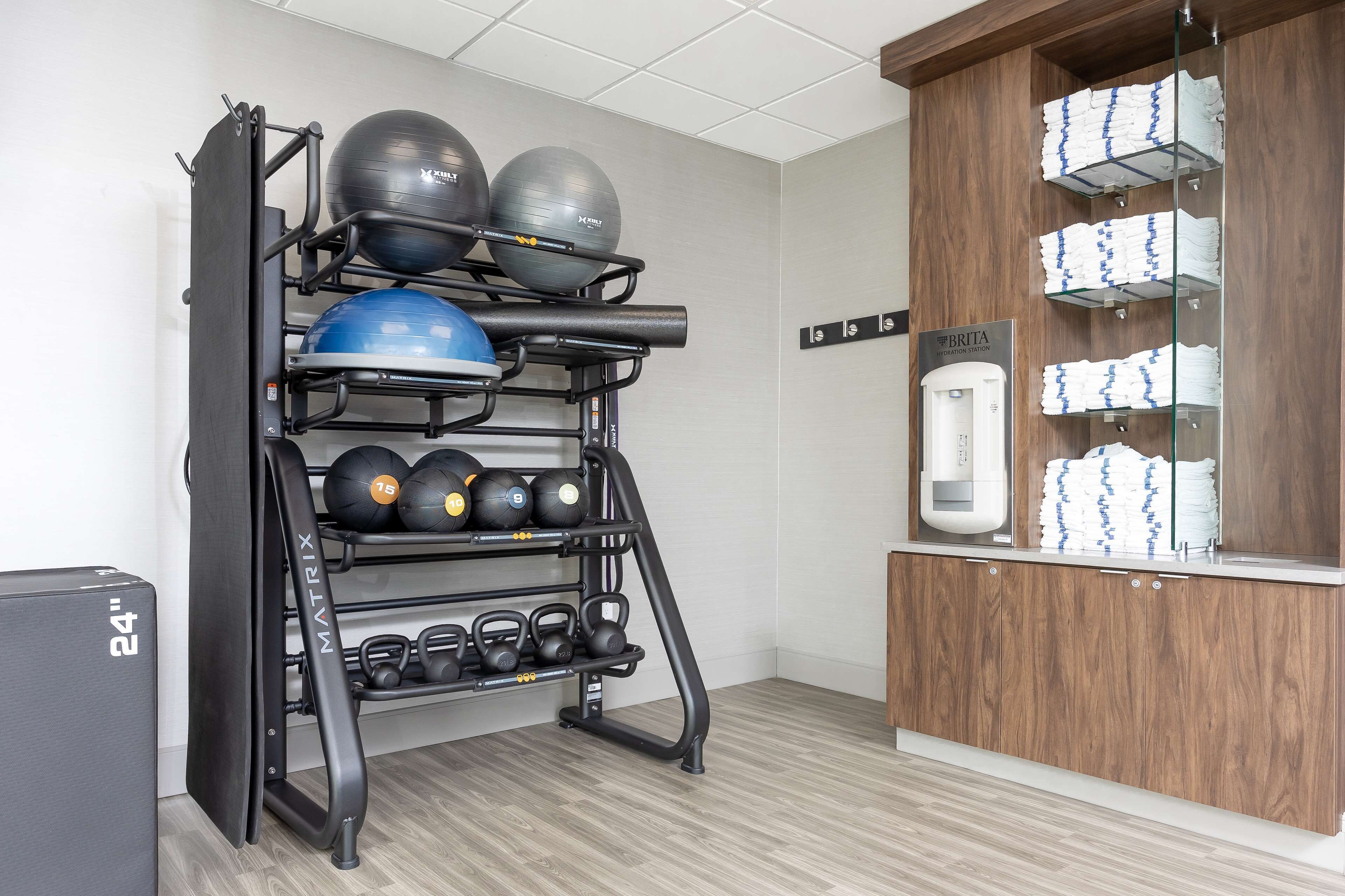 A gym with a wall-mounted rack holding various exercise balls and weights, a water dispenser, and neatly stacked towels.