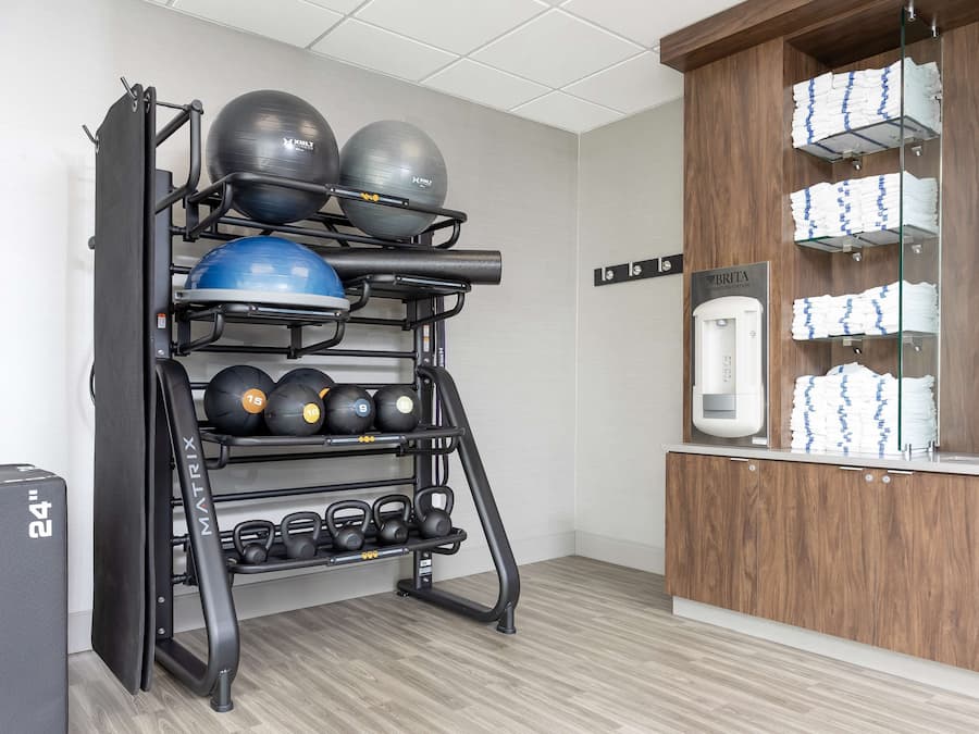 Uma academia com um suporte fixado na parede que contém bolas de exercício e pesos variados, um bebedouro e toalhas empilhadas de forma organizada.