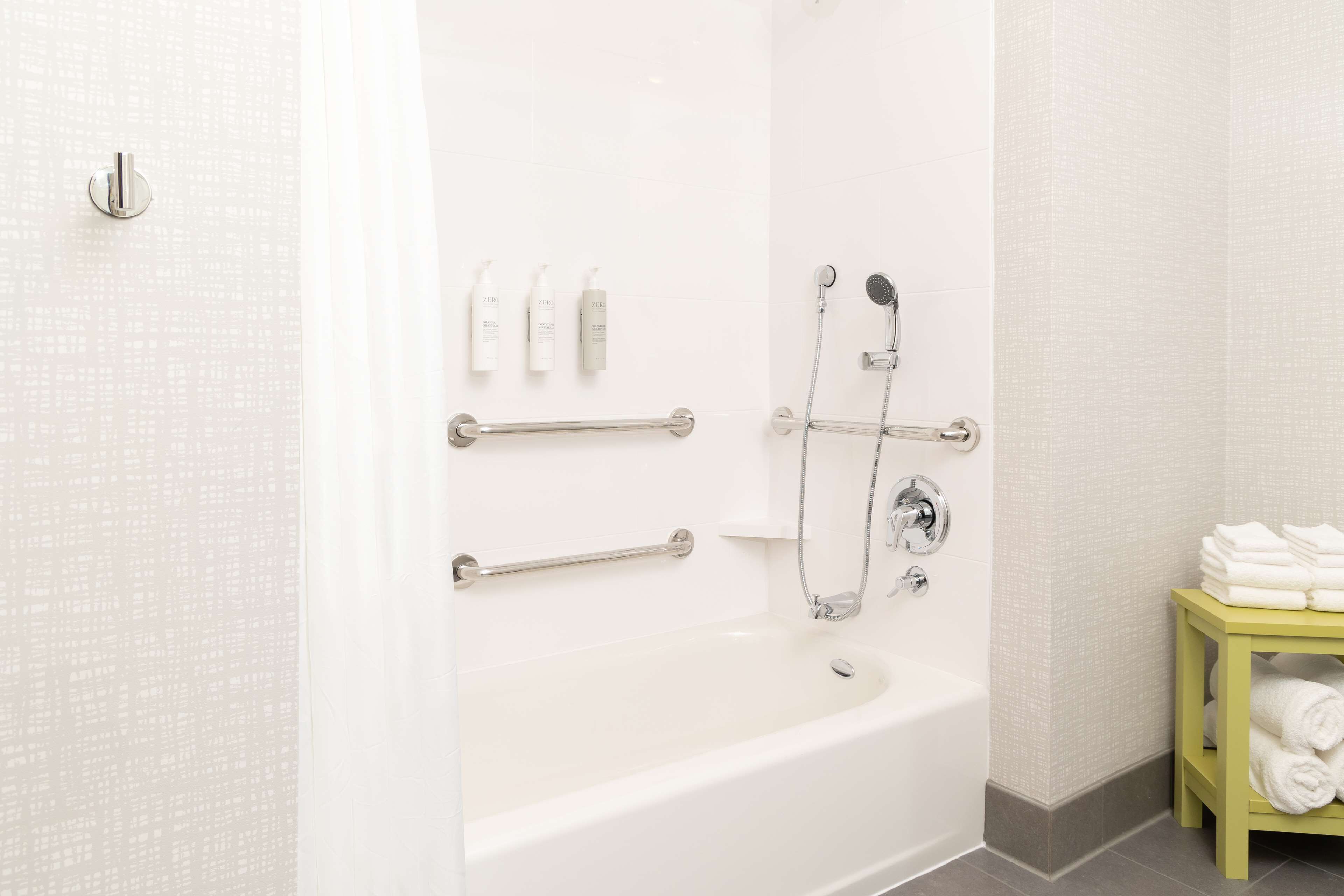 A bathroom with a white bathtub, grab bars, a showerhead, and a shelf with towels.