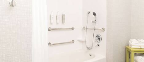 A bathroom with a white bathtub, grab bars, a showerhead, and a shelf with towels.