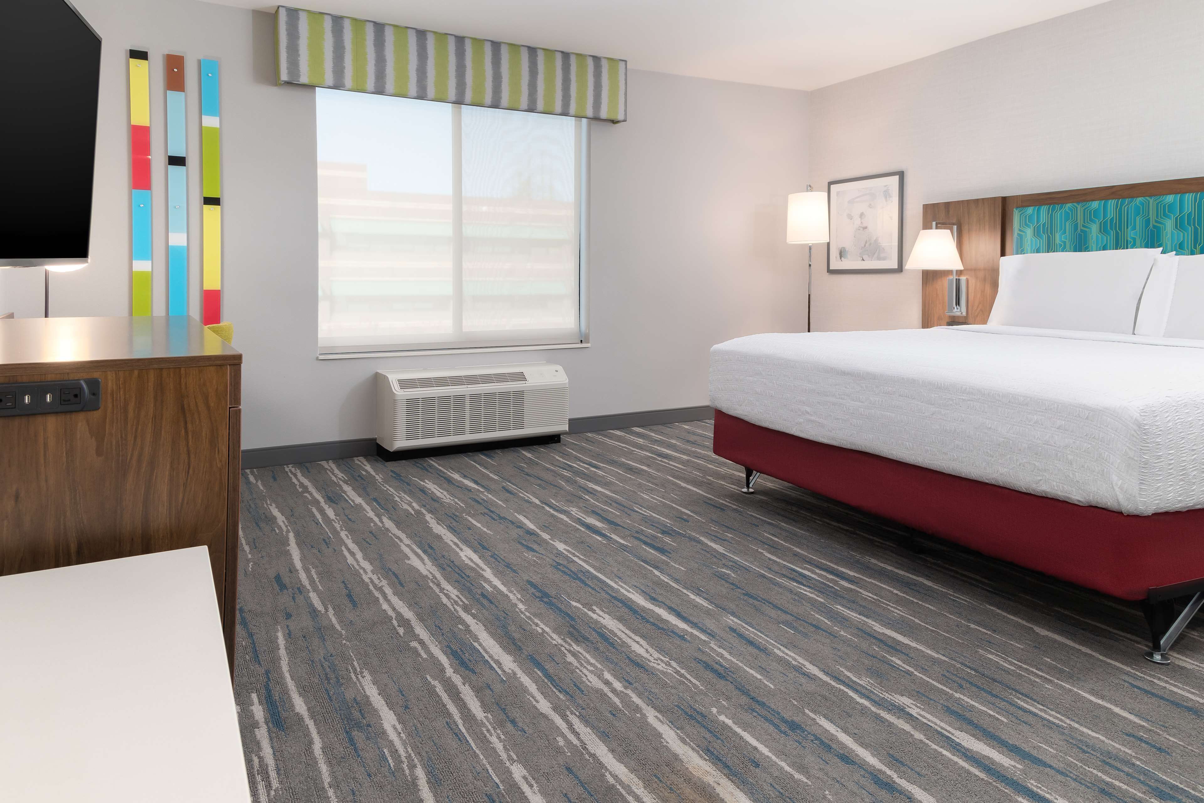 A hotel room with a bed, a desk, a TV, a window with blinds, a wall-mounted air conditioner, and a patterned carpet.