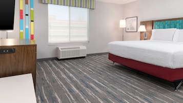 A hotel room with a bed, a desk, a TV, a window with blinds, a wall-mounted air conditioner, and a patterned carpet.