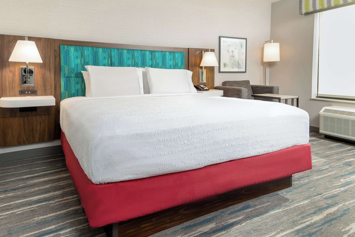 A hotel room with a large bed, a red upholstered headboard, two bedside tables with lamps, a small desk, and a large window.