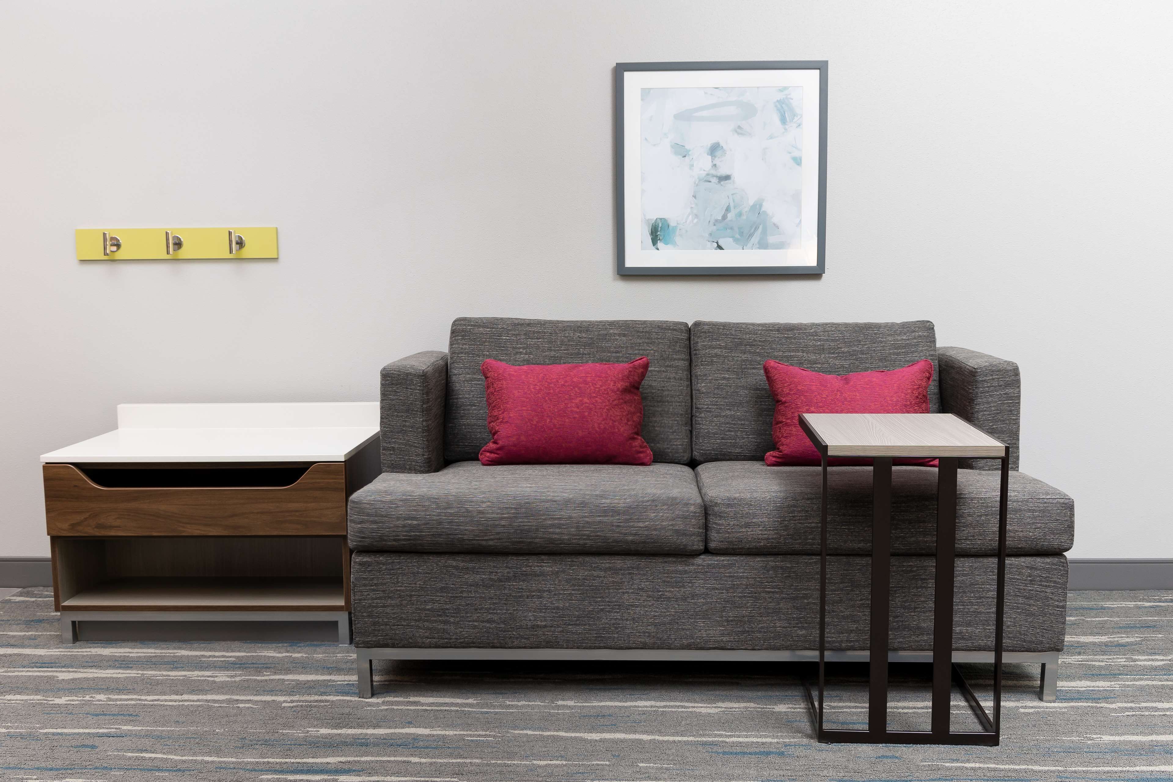 A modern living room with a grey sofa, two red cushions, a small side table, and a framed abstract painting on the wall.