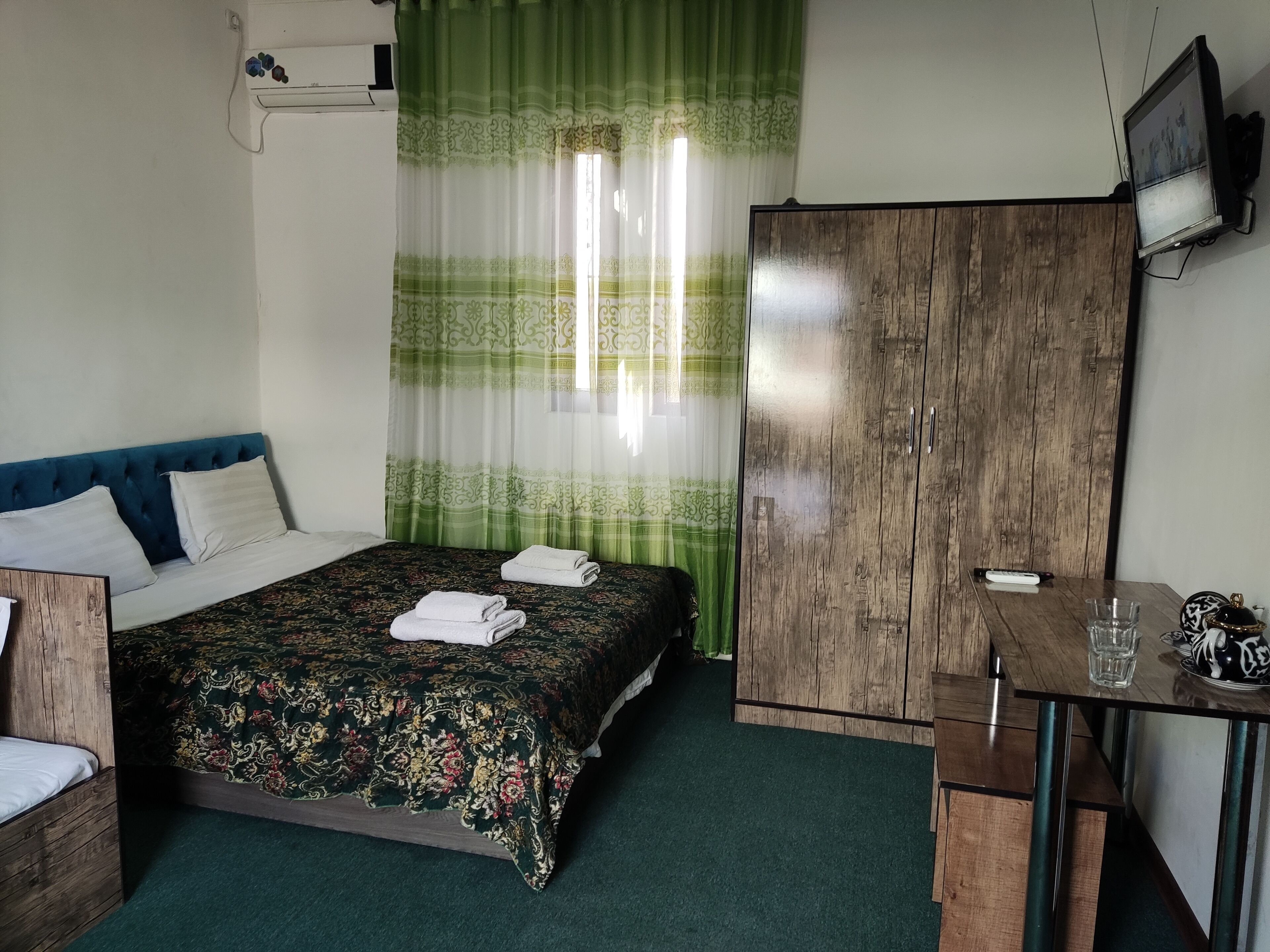 A bedroom with a bed, a wooden wardrobe, a desk with a chair, and a small table with a glass.
