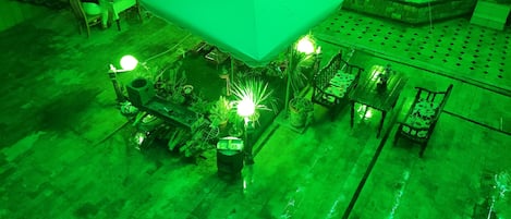 An outdoor seating area with tables and chairs, surrounded by potted plants and a green-lit canopy.