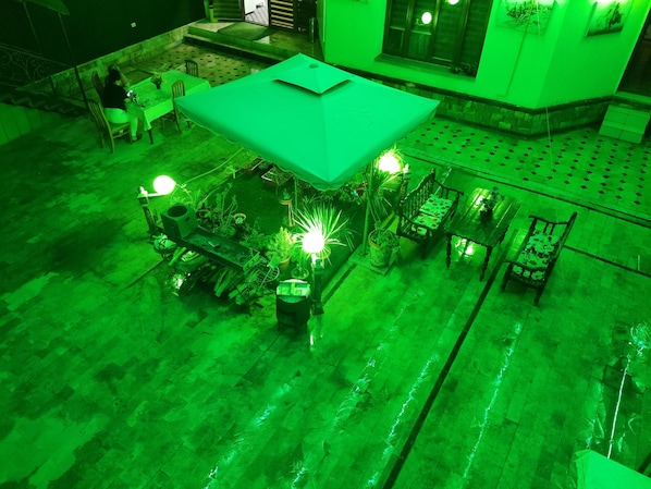 An outdoor seating area with tables and chairs, surrounded by potted plants and a green-lit canopy.
