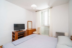 Une chambre à coucher avec un lit, une télévision, une commode, une chaise et une fenêtre avec des rideaux.