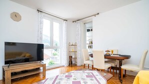 Una sala de estar con televisor, una mesa de comedor con sillas, un sillón amarillo, una mesa de centro de madera y un ventanal con cortinas.