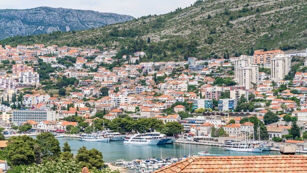 Un pueblo costero con edificios muy juntos, un puerto deportivo con barcos y un paisaje montañoso de fondo.