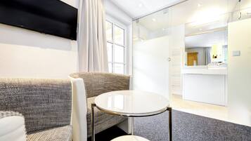 A modern hotel room with a sofa, a small round table, a flat-screen TV mounted on the wall, and a glass partition separating the room from the bathroom area.