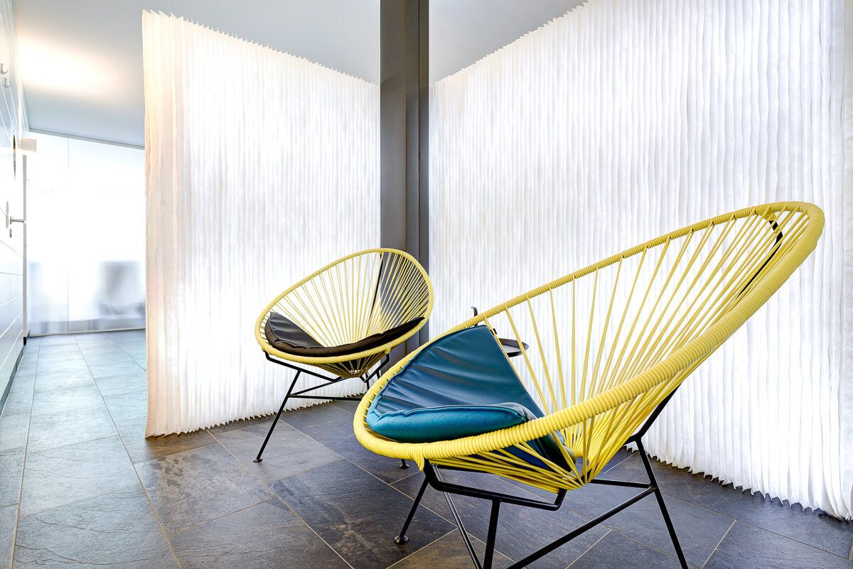 Two yellow wicker chairs with blue cushions in a room with dark tiled flooring and white curtains.