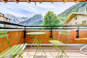 Un balcón con piso de madera, sillas de metal verdes, una mesa redonda y vistas a montañas y un árbol.