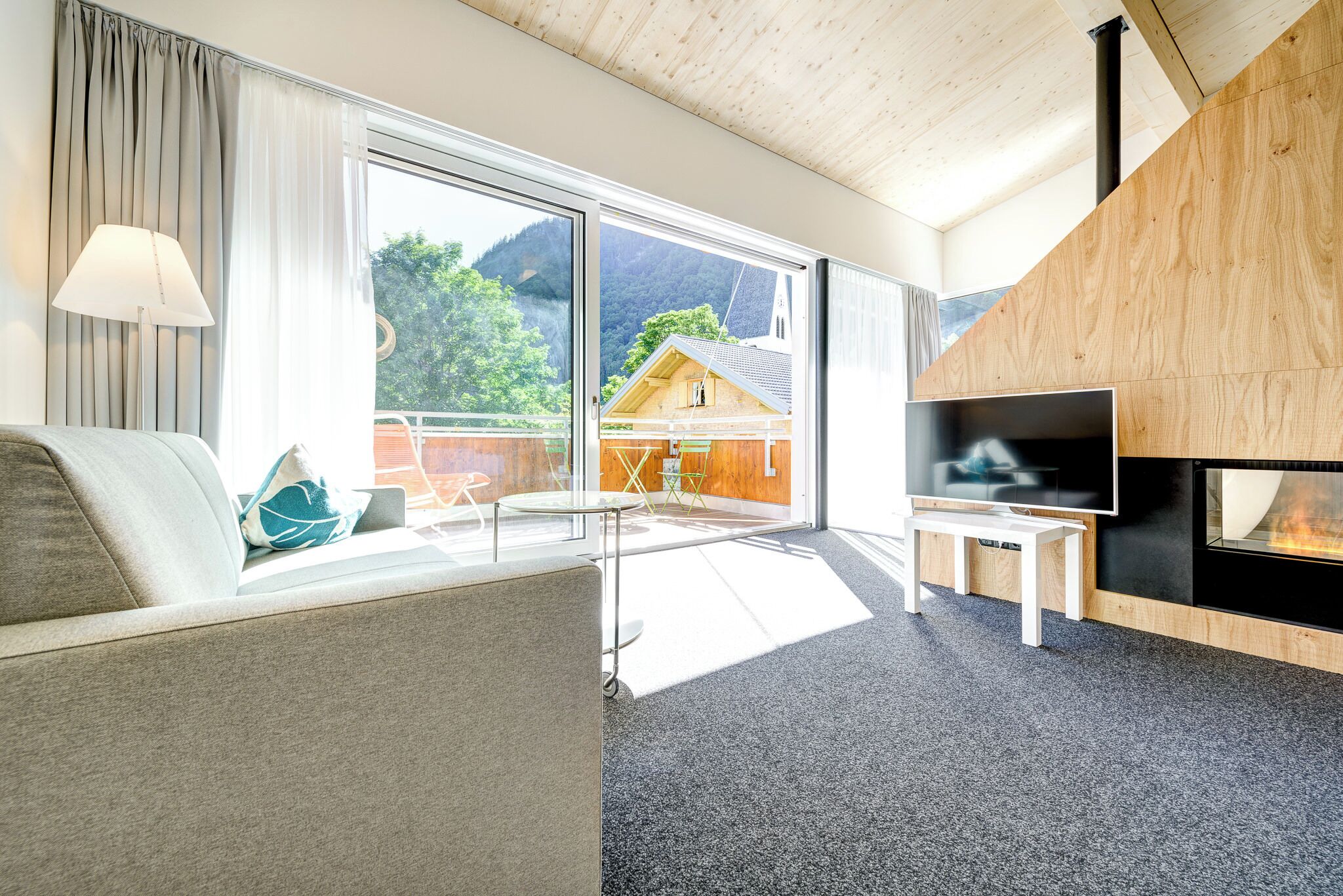 Un salon moderne avec un canapé, un téléviseur, une cheminée et une vue sur une montagne et une maison, le tout visible à travers de grandes fenêtres.