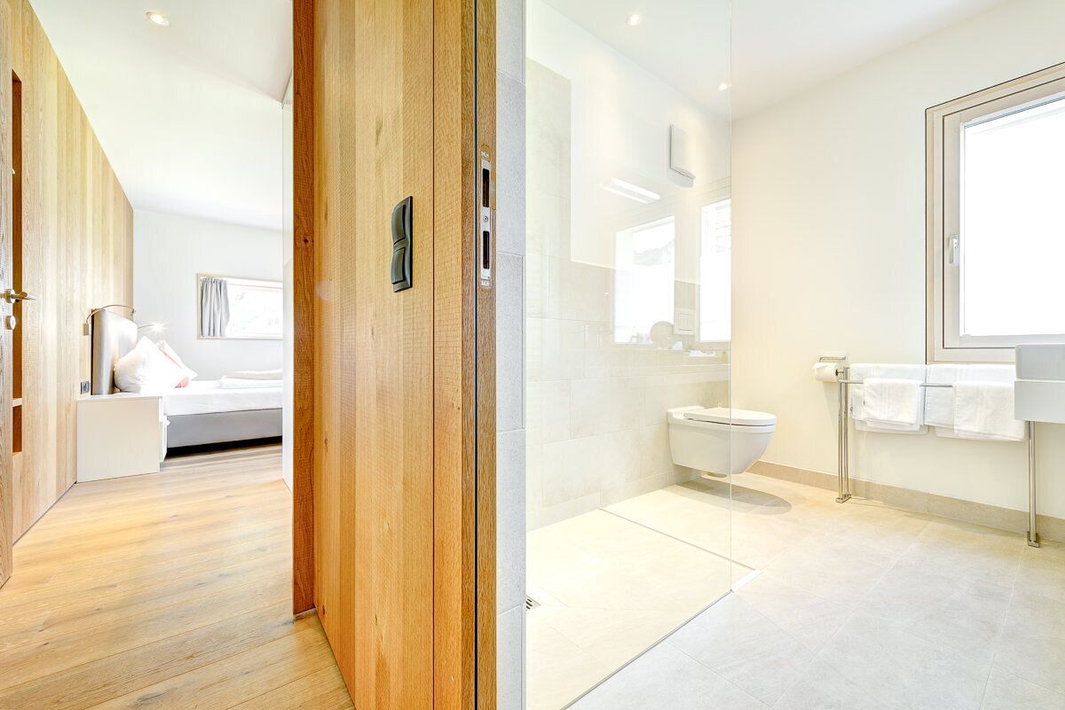 A modern hotel room with a wooden floor, a bed with white bedding, a glass shower enclosure, a toilet, and a large window.
