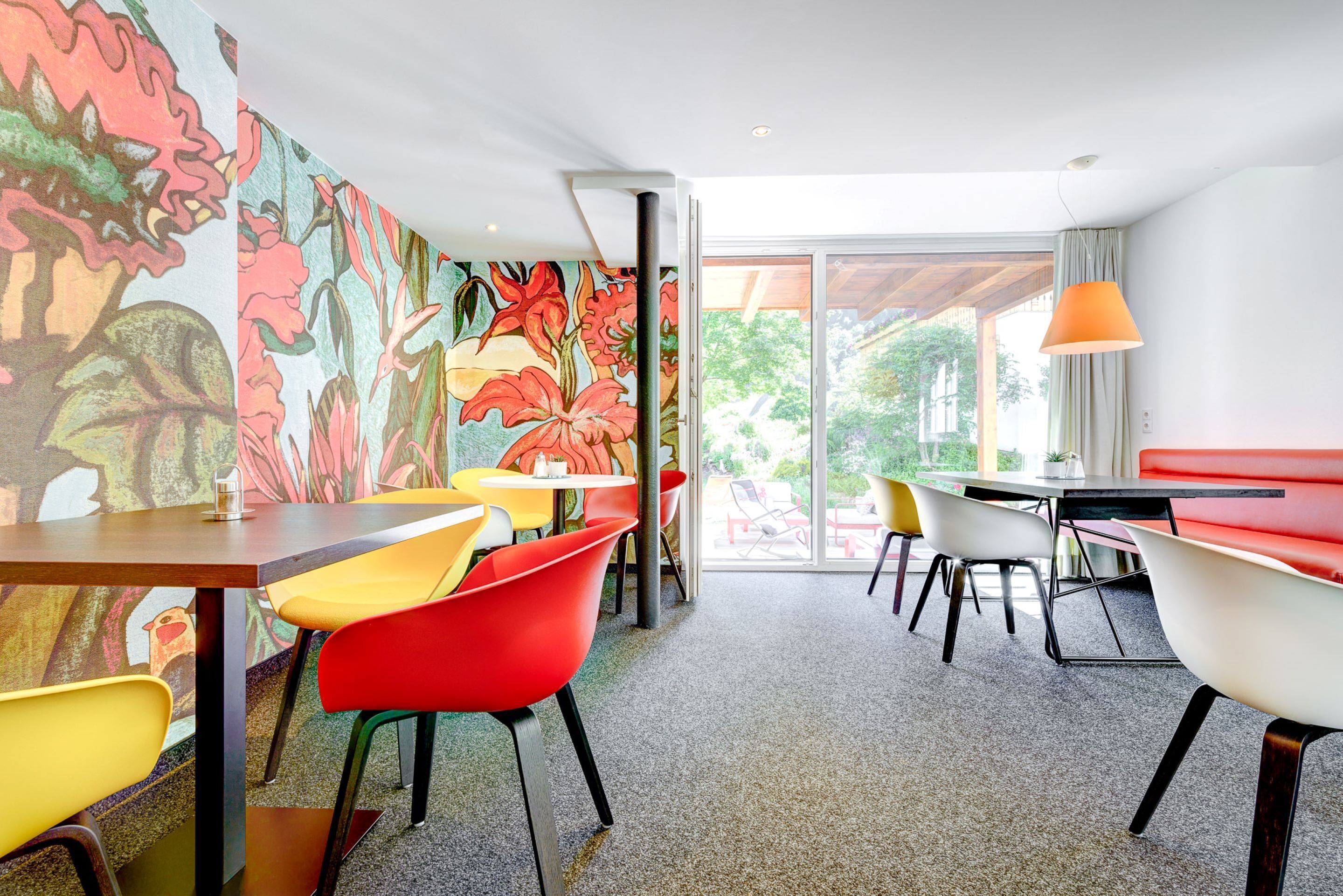 Een eetruimte met kleurrijke stoelen en tafels, een grote muurschildering en uitzicht op het buitengebied door grote ramen.
