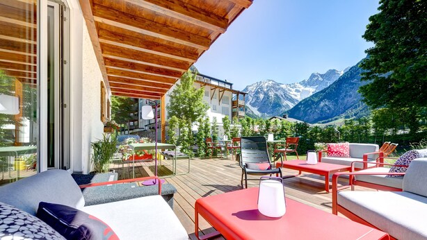 Un terrazzo in legno con mobili da esterno, vista sulle montagne e un edificio con balconi.