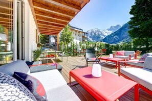 Una terraza de madera con mobiliario exterior, vistas a la montaña y un edificio con balcones.