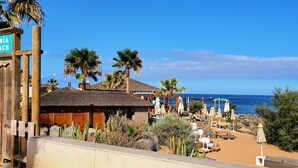 A beachside resort with a sign reading "Bahia Beach," wooden structures, palm trees, and a view of the ocean. - Luxury Oceanfront Apartment with beautiful Seaview, WIFI, Pool (Palm-Mar)