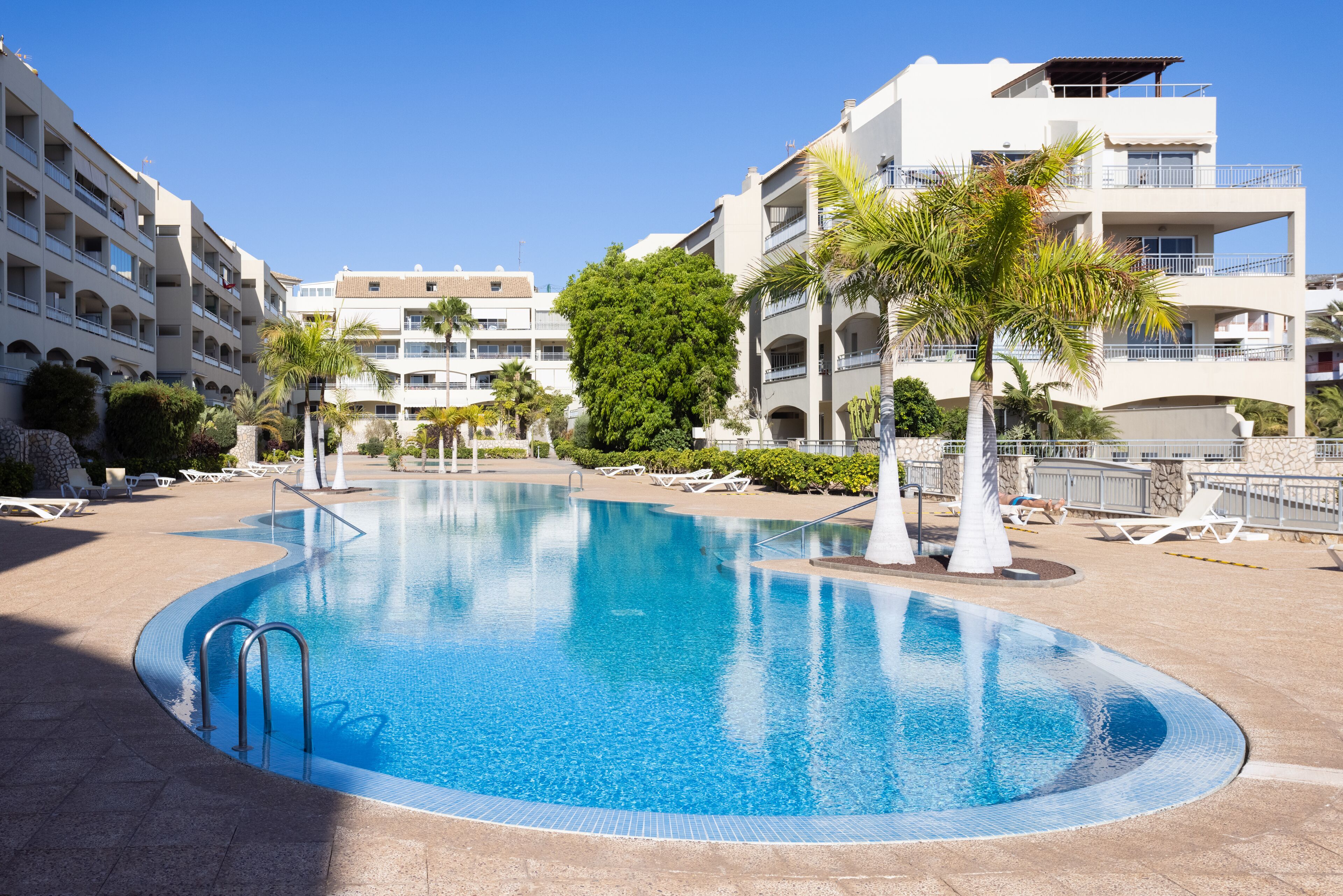 A swimming pool surrounded by lounge chairs and palm trees, with multi-story buildings in the background.
