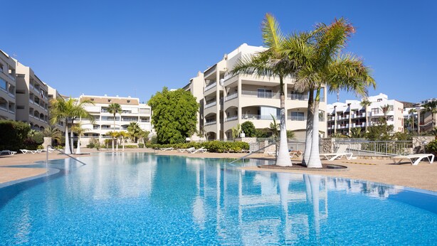 A clear blue pool surrounded by white buildings with balconies, palm trees, and lounge chairs. - Luxury Oceanfront Apartment with beautiful Seaview, WIFI, Pool (Palm-Mar)