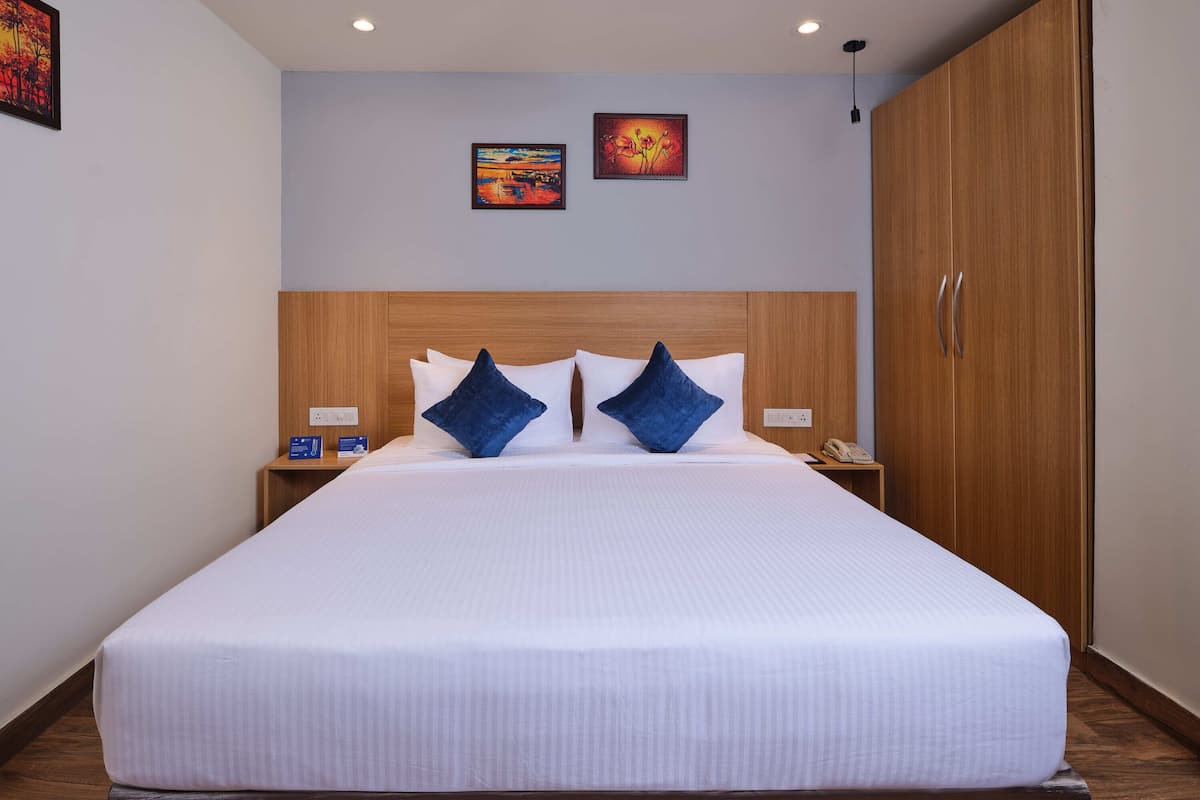 Habitación de hotel con una cama grande, dos almohadas azules, un cabecero de madera, una mesita de noche con un teléfono y dos cuadros enmarcados en la pared.