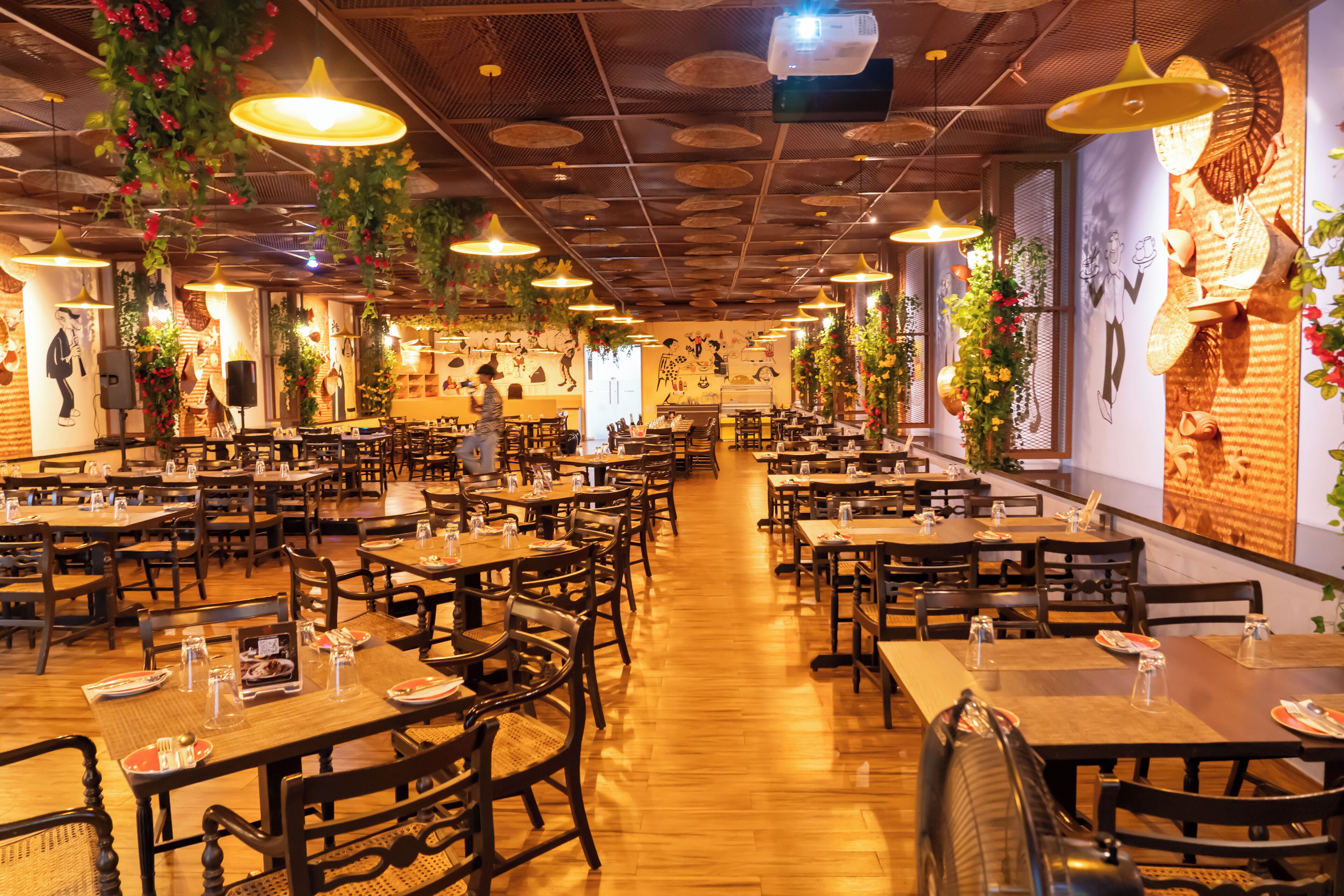 A restaurant with multiple tables and chairs, hanging plants, and a patterned ceiling.