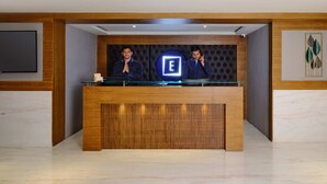 A reception desk with a logo, two individuals standing behind it, and a modern interior design. - Essentia Premier Hotel Chennai OMR (Chennai)