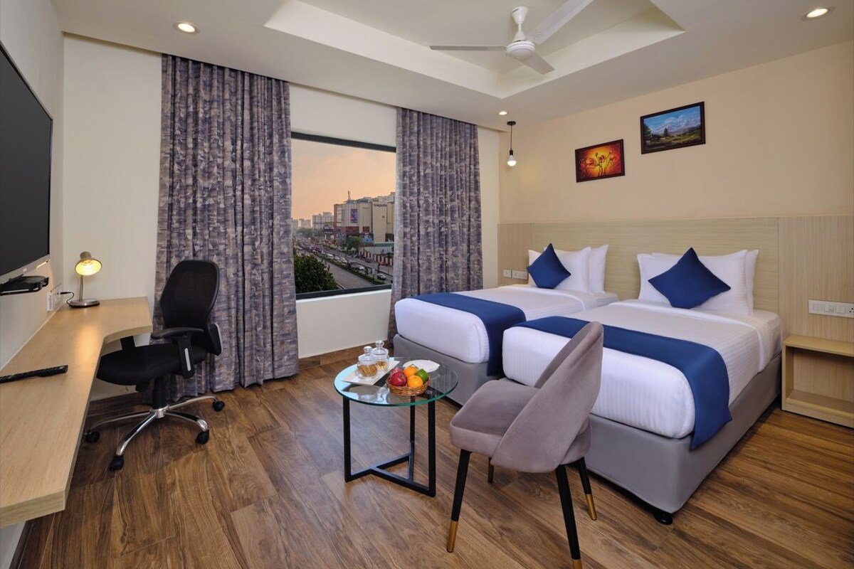 A hotel room with a desk, two beds, a chair, a small table with fruit, and a city view.