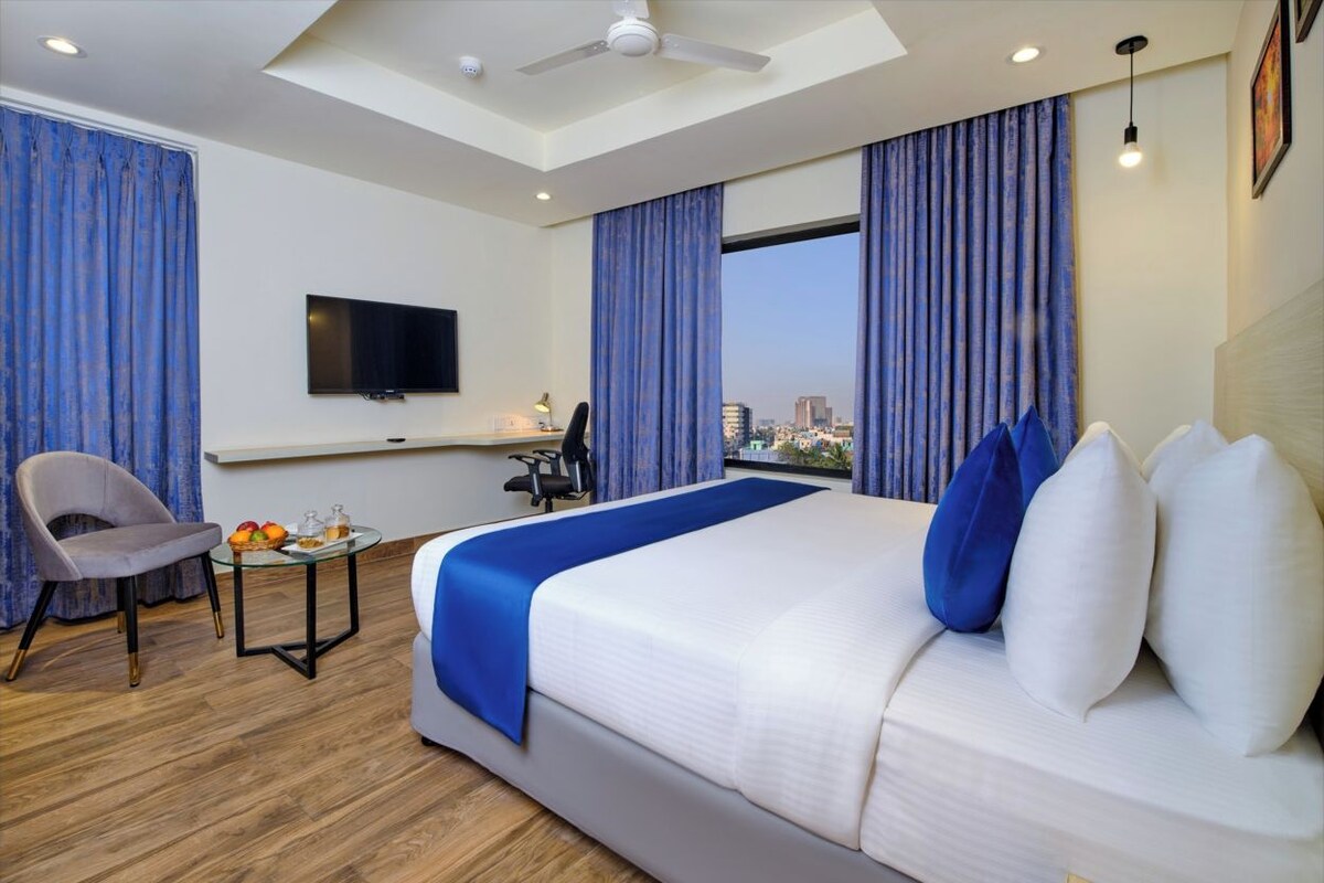 Habitación de hotel moderna con una cama grande, un escritorio con silla, un televisor y vistas a la ciudad a través de las ventanas.