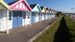 A row of colorful beach huts with white doors and windows, arranged along a paved path beside a grassy area. - Deer Leap House (Dorchester)