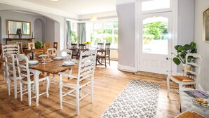 A dining room with a wooden table and white chairs, large windows, and a view of greenery outside. - Deer Leap House (Dorchester)