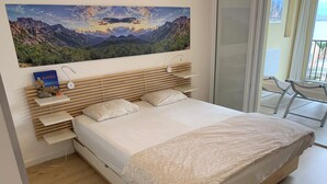 A modern bedroom with a wooden bed frame, white bedding, and a landscape painting above the bed. - Family Vacation Apartment with Panoramic Sea View (Calvi)