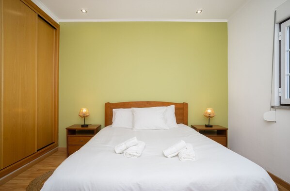 A neatly made bed with white linens and towels, two bedside lamps, and a wooden wardrobe.