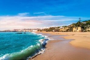 A coastal beach with clear blue waters, a sandy shore, and a town in the background.