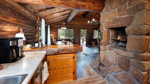 Ein rustikales Blockhaus mit Kamin aus Naturstein, Holzbalken und einer Küchenzeile mit Kaffeemaschine und Weinflasche.