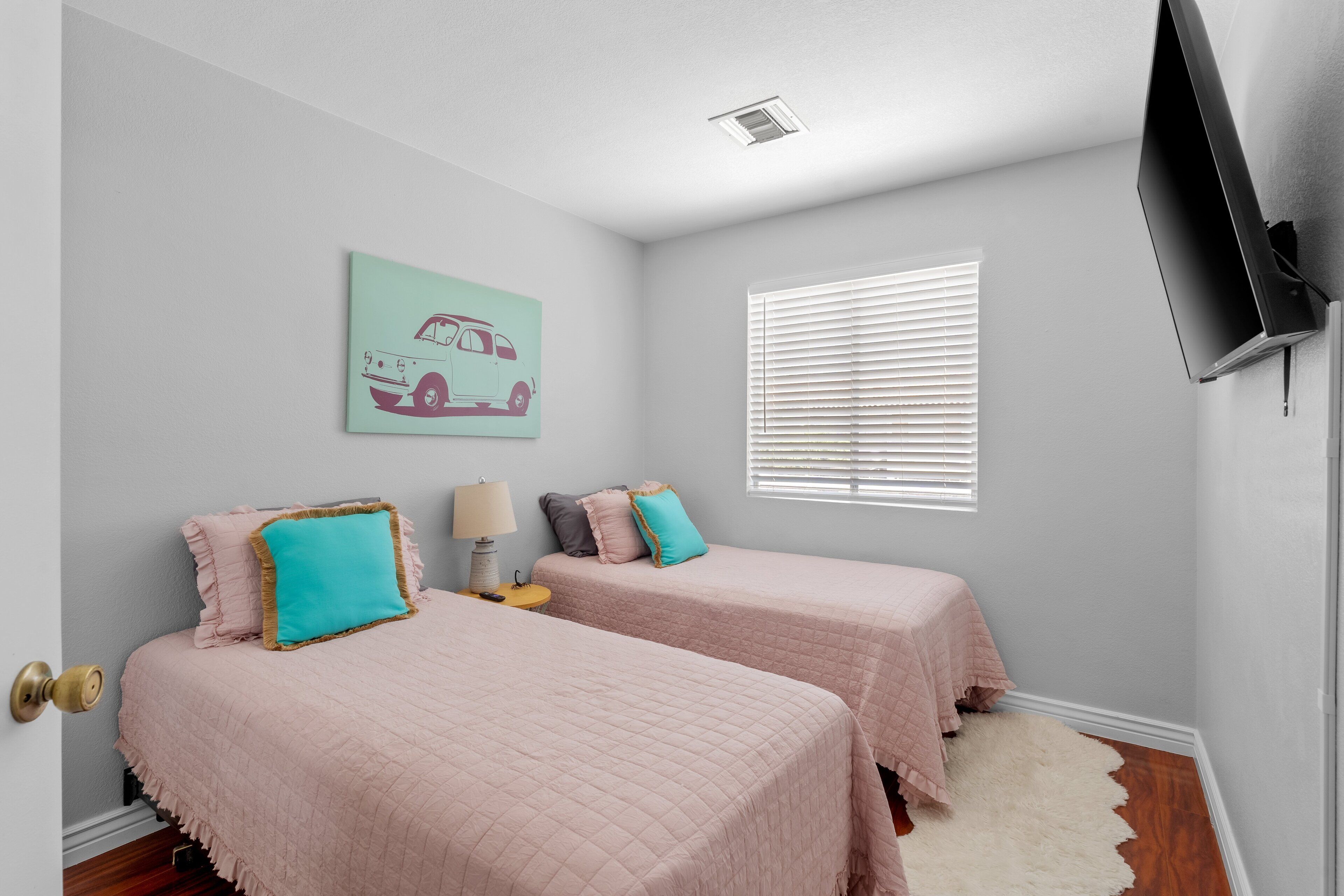 A bedroom with two beds, a TV, a window with blinds, a door, and a wall-mounted artwork.