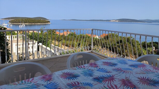 A balcony with a table covered in a floral tablecloth, overlooking a coastal view with boats and a distant island.