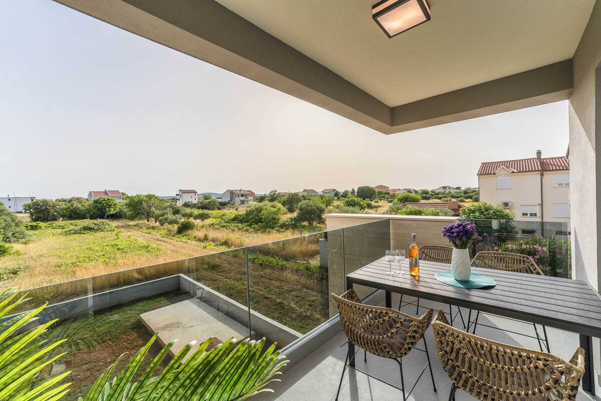 Uma varanda com barreira de vidro, cadeiras de vime, uma mesa com vaso e garrafa, e vista para uma área residencial com vegetação e casas.
